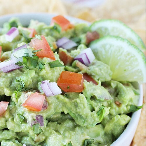 Guacamole in a white bowl topped with chopped red onion, tomato, and cilantro.