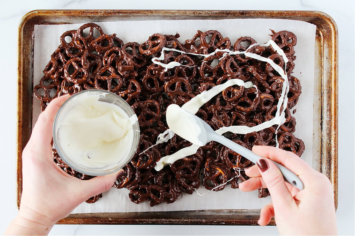 A woman using a spatula to drizzle melted white chocolate over chocolate covered pretzels.