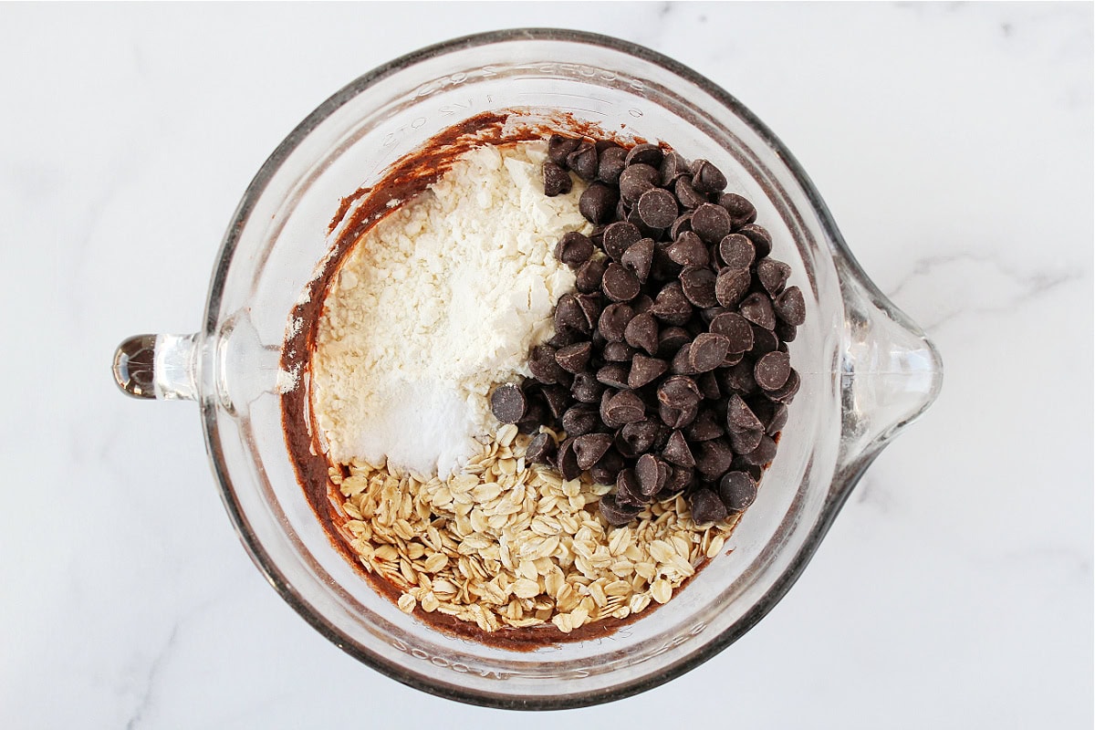 Dark chocolate chips, rolled oats, and flour on top of chocolate batter in a bowl.
