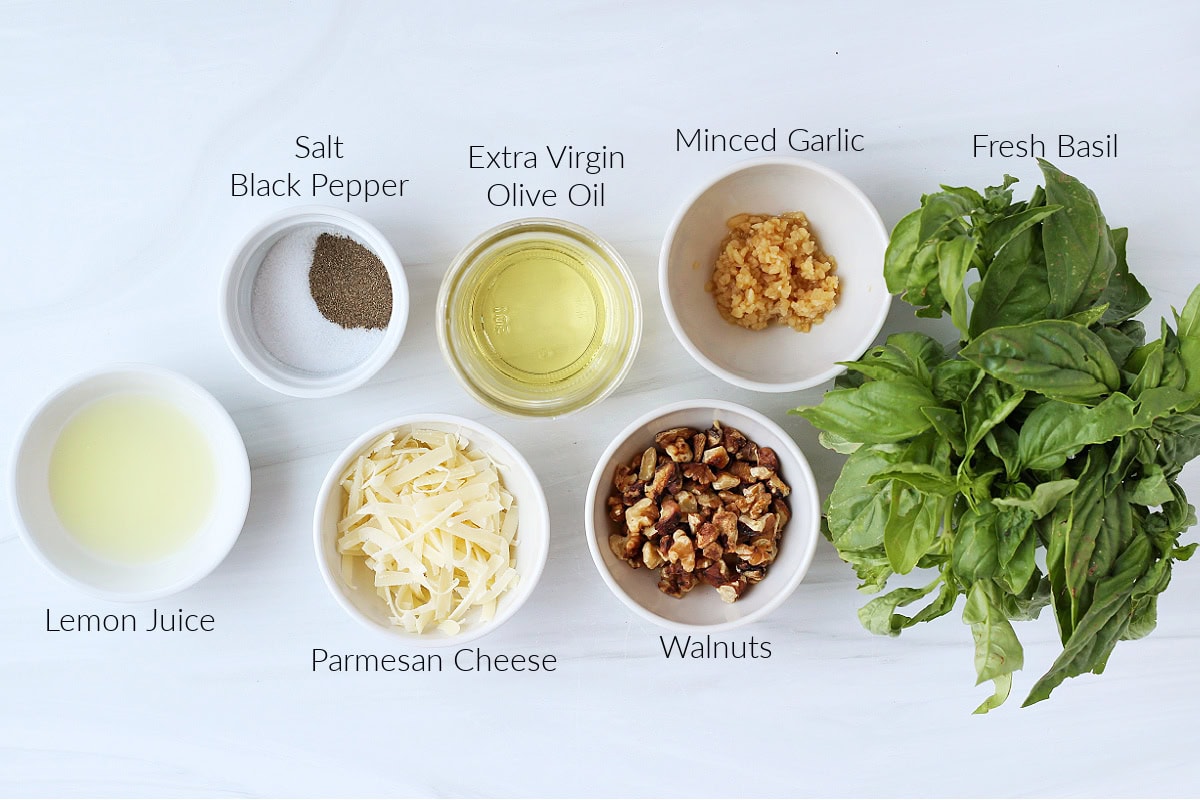 Labeled ingredients for homemade basil walnut pesto on a white marble countertop.
