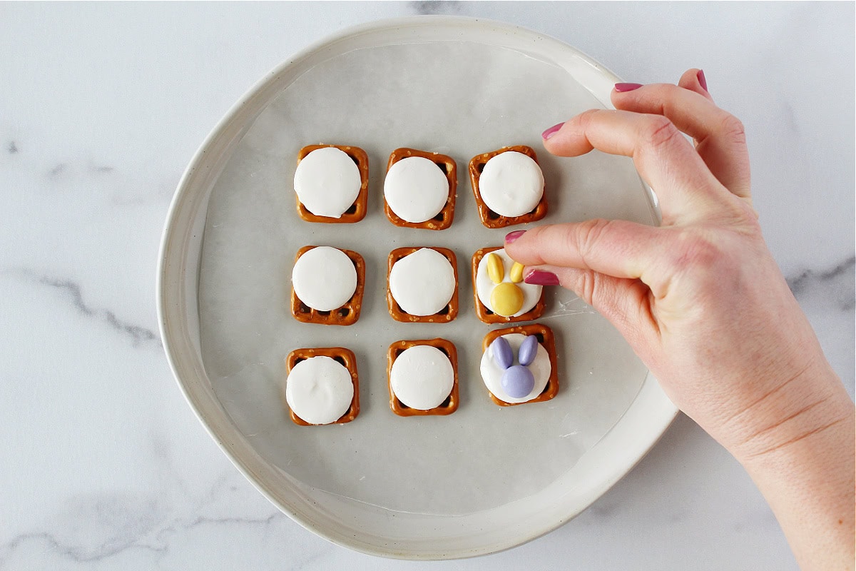 A woman placing a halved M&M on a melty white candy wafer on a pretzel.