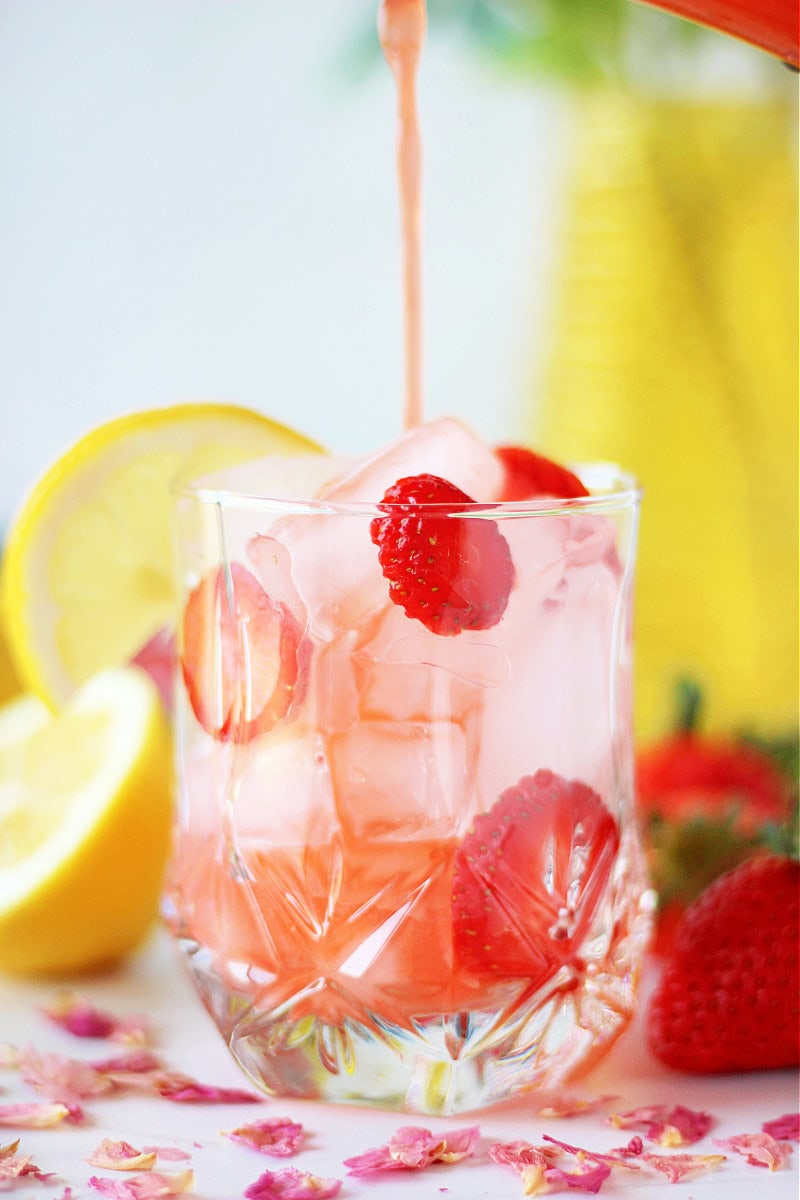 Pouring strawberry lemonade into a glass of ice cubes and strawberry slices.
