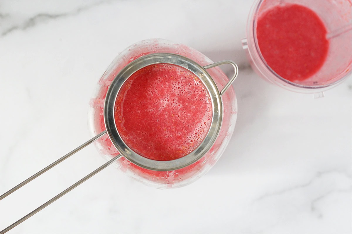 Small metal strainer over a glass jar straining raspberry puree.