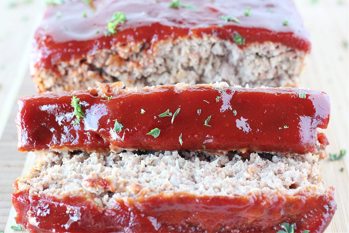 Slices of ketchup sauce topped meatloaf on a cutting board.