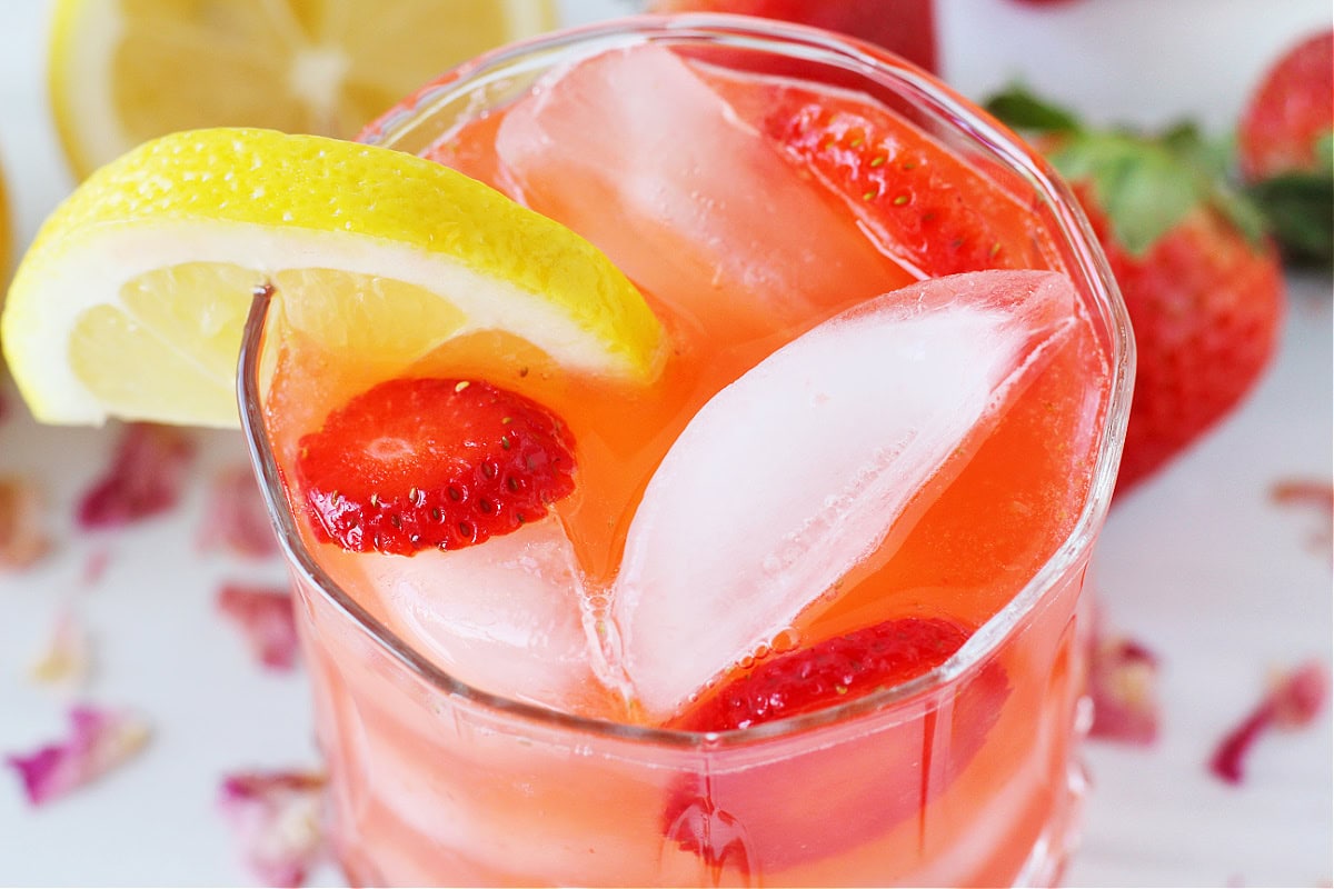 A lemon slice and sliced strawberries in a glass of iced strawberry lemonade.