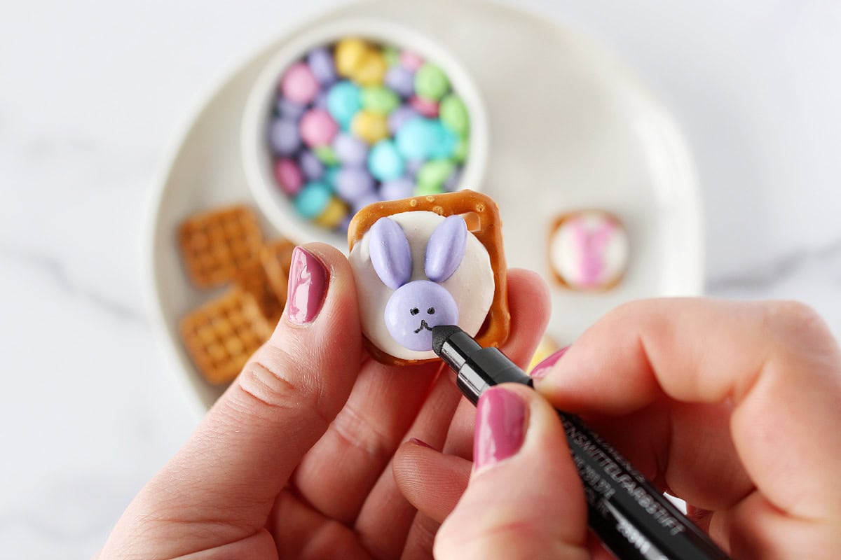A woman using a black edible marker to draw a bunny face on Easter Bunny Pretzels.