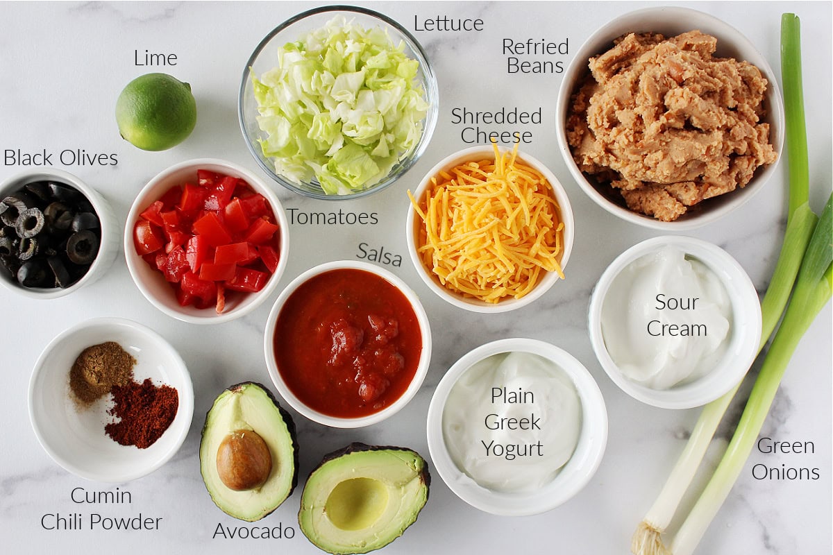 Labeled ingredients in bowls for 9 layer Mexican dip on a white marble countertop.