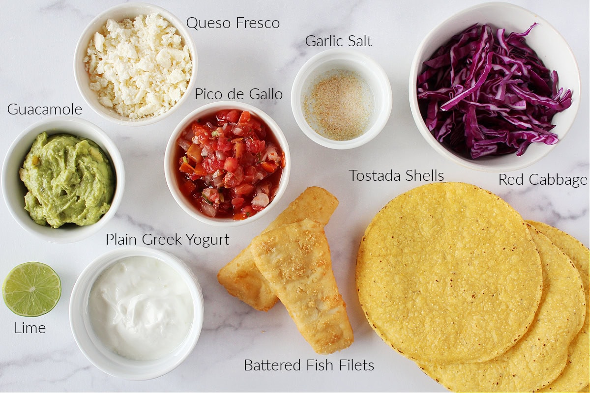 Labeled ingredients for fish tostadas with cilantro lime crema on a white marble countertop.