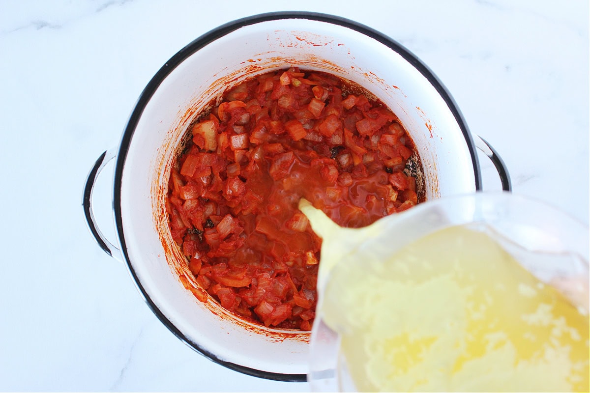Pouring broth into sauteed onions and tomato paste in a soup pot.