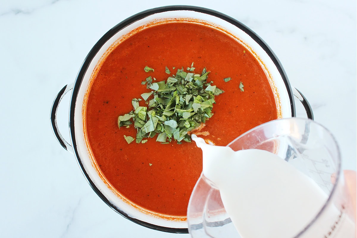 Pouring half and half into a pot of pureed tomato soup topped with chopped basil.