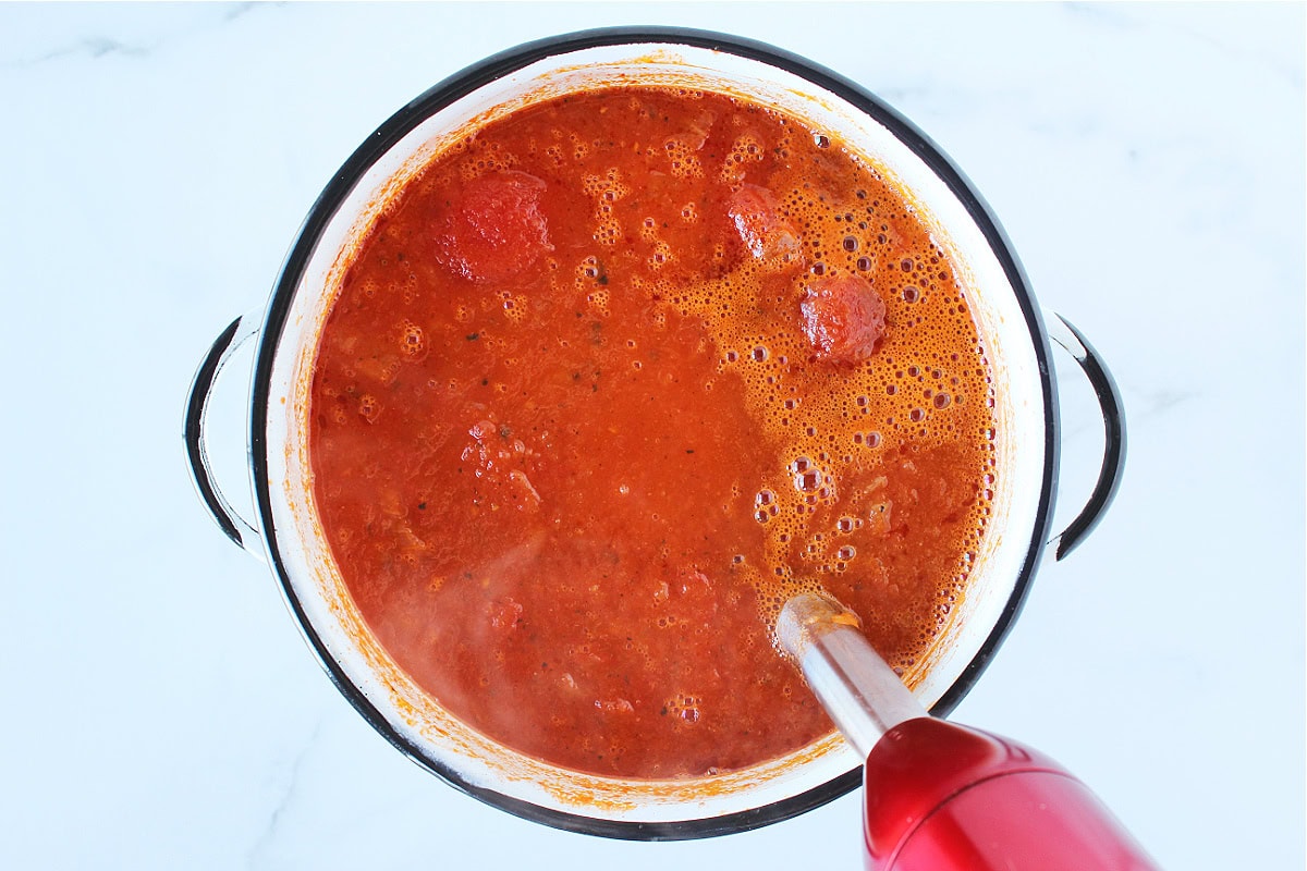 An immersion blender blending tomato soup in a white soup pot.