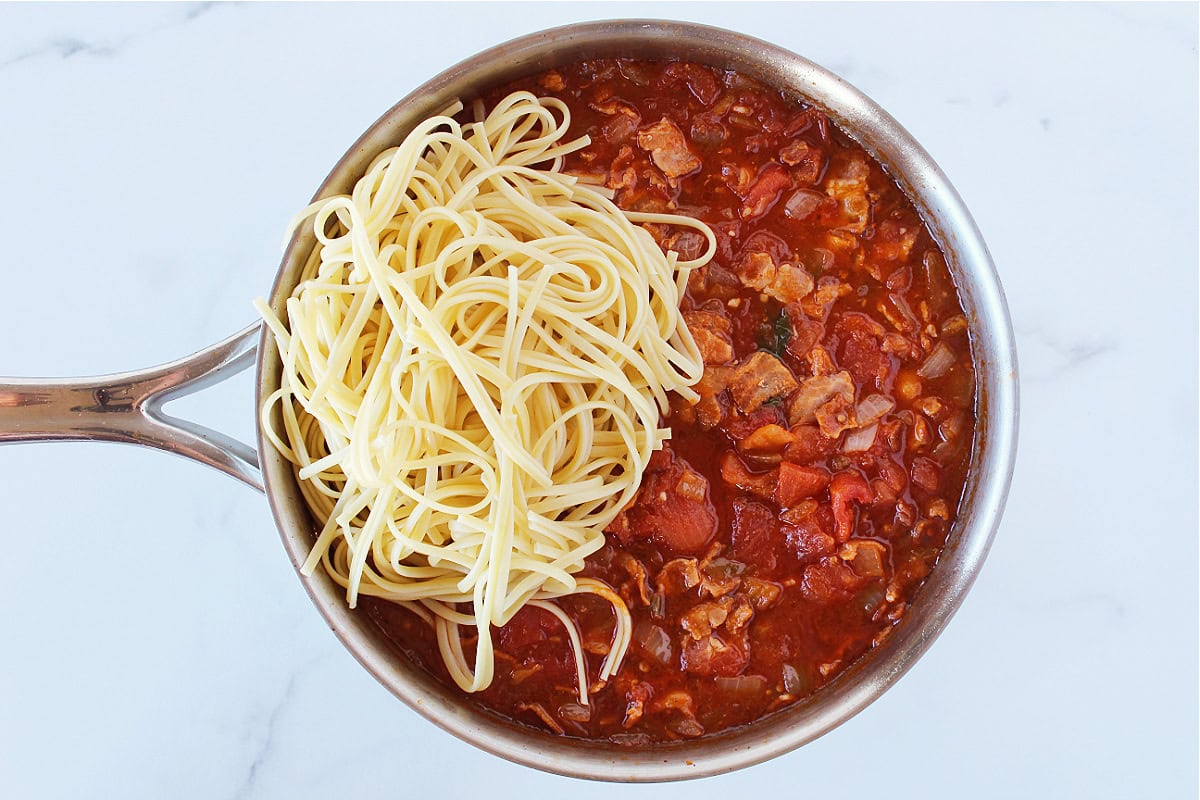 Linguine pasta sitting atop a stewed tomatoes and bacon sauce in a large sauté pan.