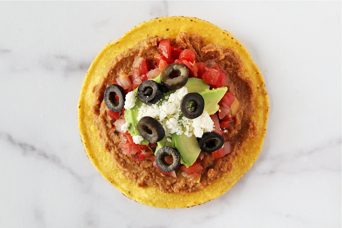 Sliced black olives, queso fresco, sliced avocado, pico de gallo, and refried beans on a tostada.