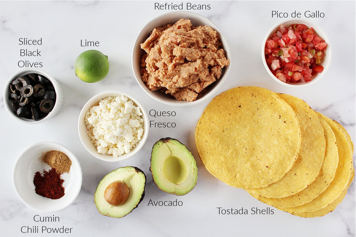 Labeled ingredients in bowls for vegetarian tostadas on a white marble countertop.