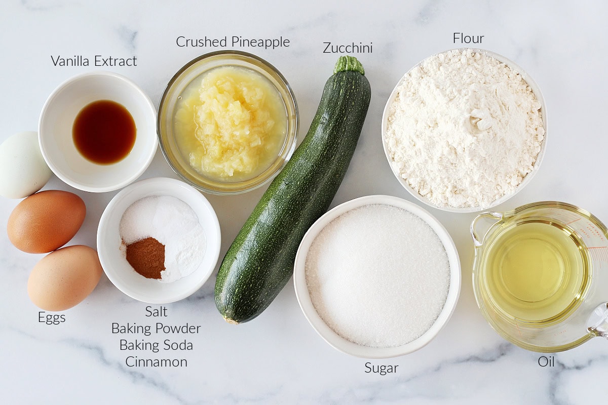 Labeled ingredients for pineapple zucchini bread on a white marble countertop.