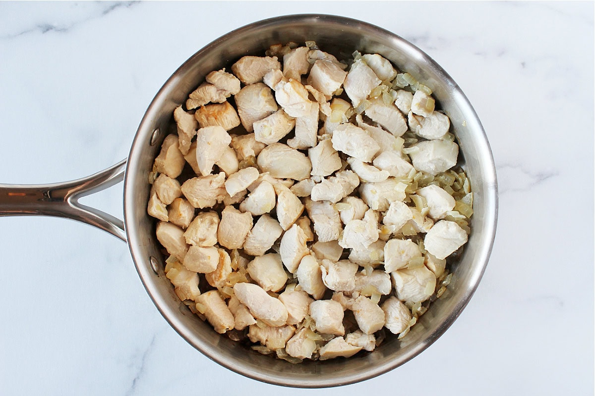 Cooked chicken breast pieces and sautéed onion in a stainless steel saute pan.