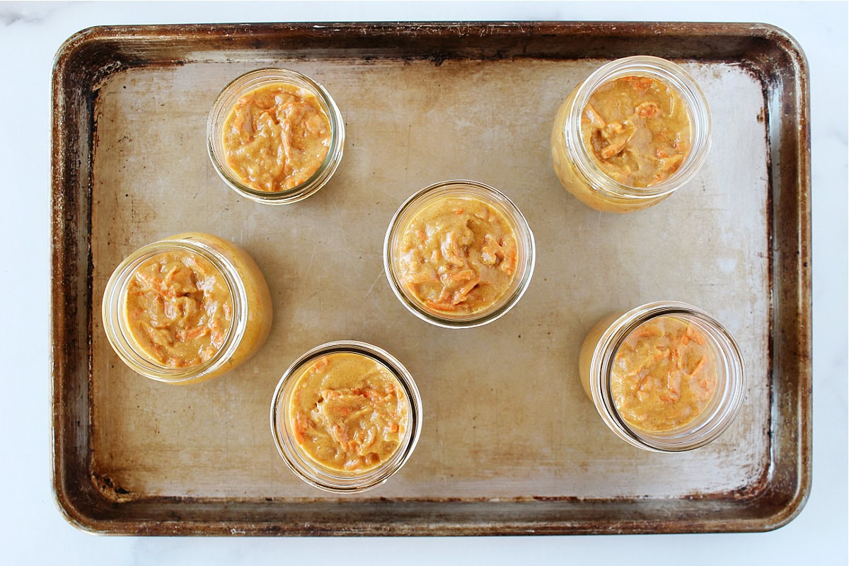 Carrot cake batter in six mason jars on a baking sheet.