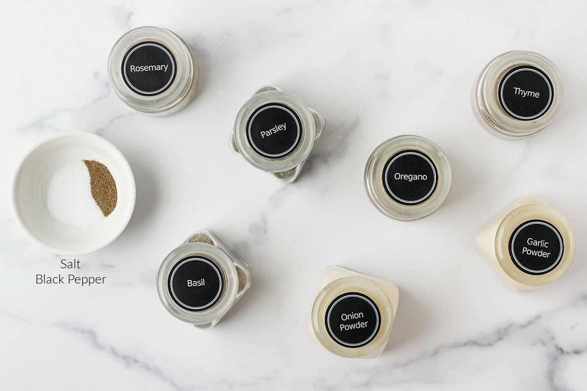 Labeled dried seasonings in jars and a bowl for Italian seasoning on a white marble countertop.