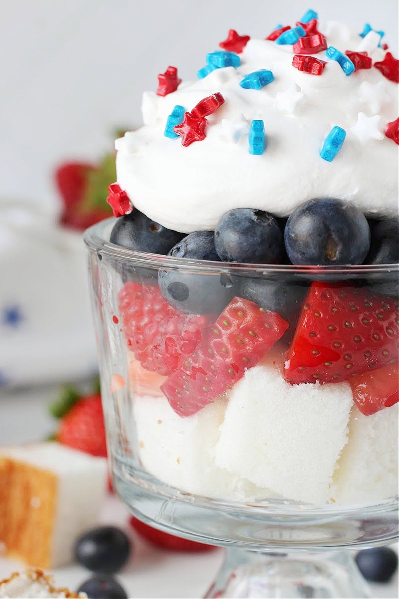 Layers of angel food cake, strawberries, blueberries, whipped cream and sprinkles in a glass.