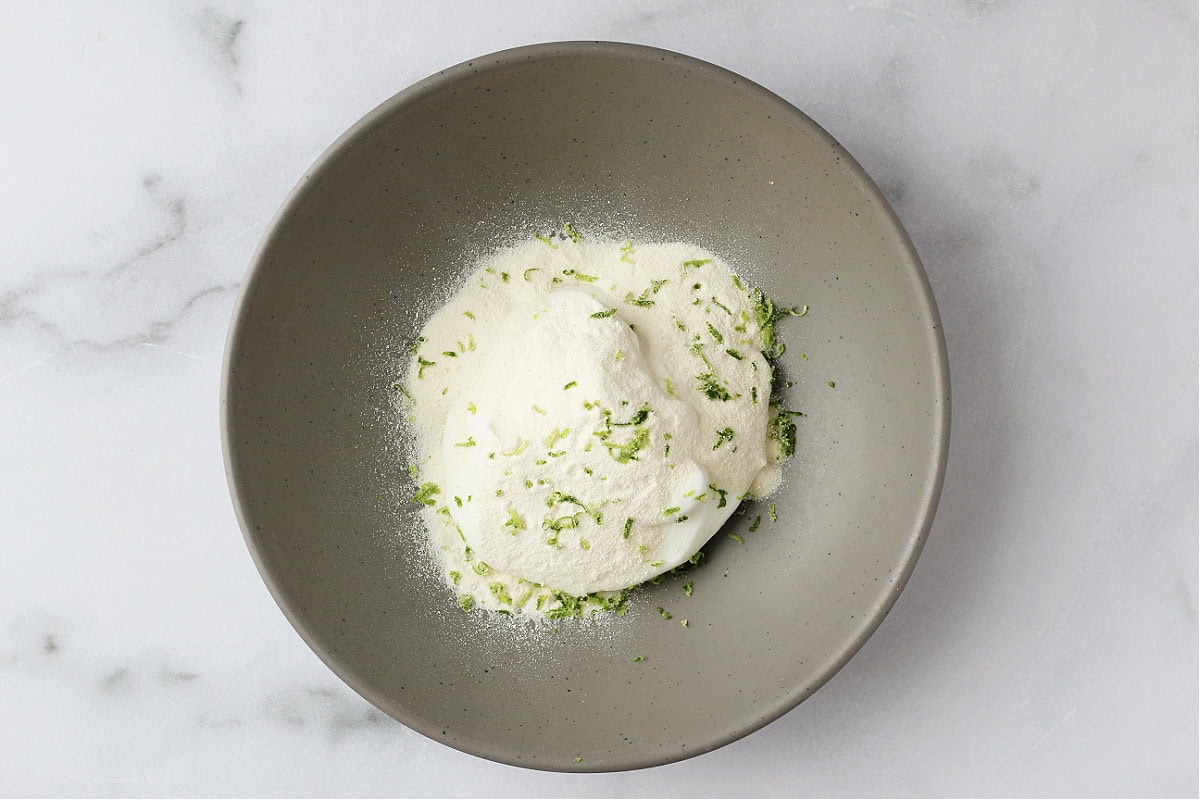 Lime zest and vanilla protein powder over plain Greek yogurt in a grey bowl.