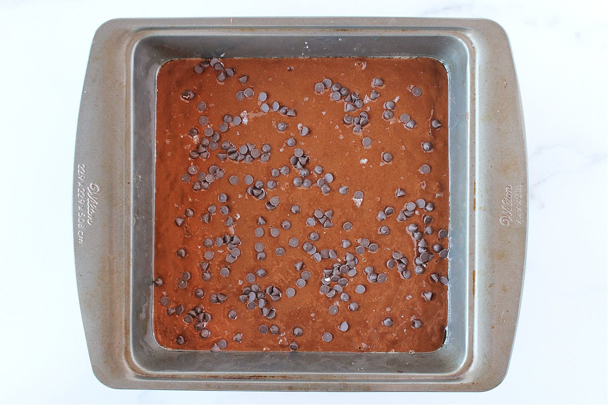 Brownie batter in a metal pan topped with mini chocolate chips and flaked salt.