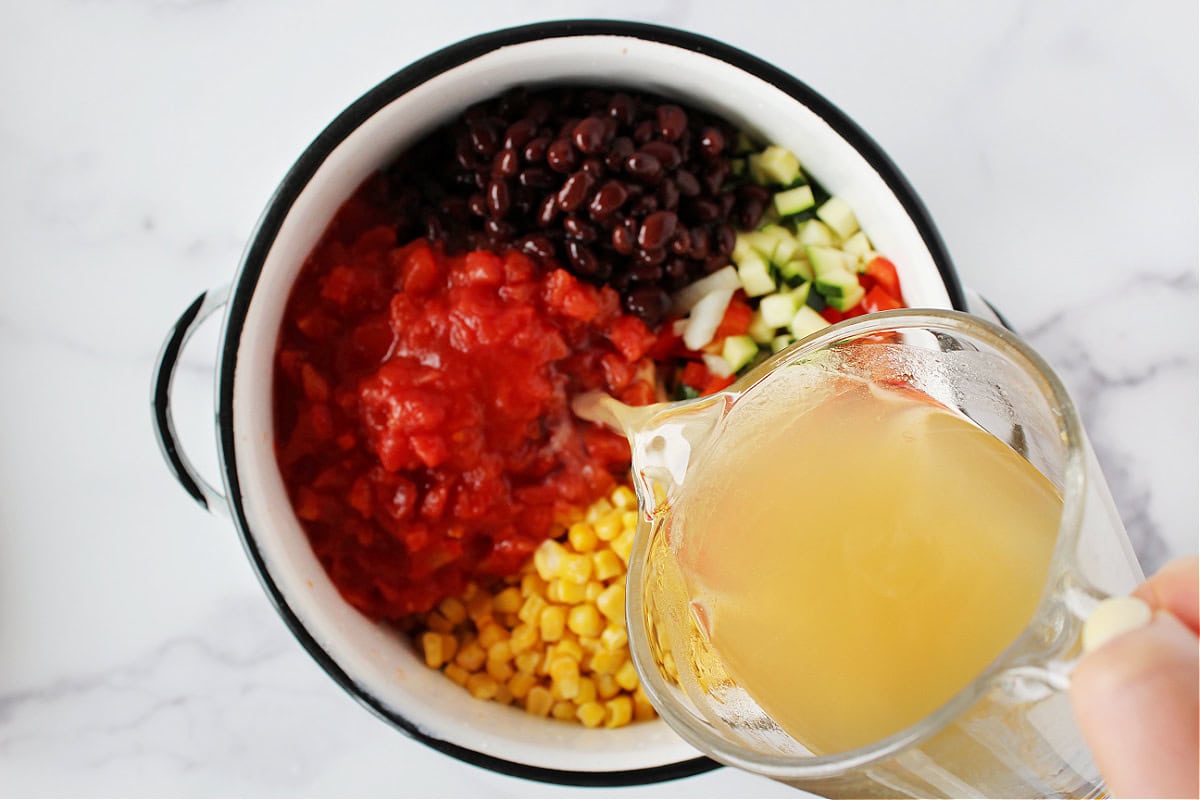 Pouring bone broth into a pot of diced tomatoes, veggies and black beans,.