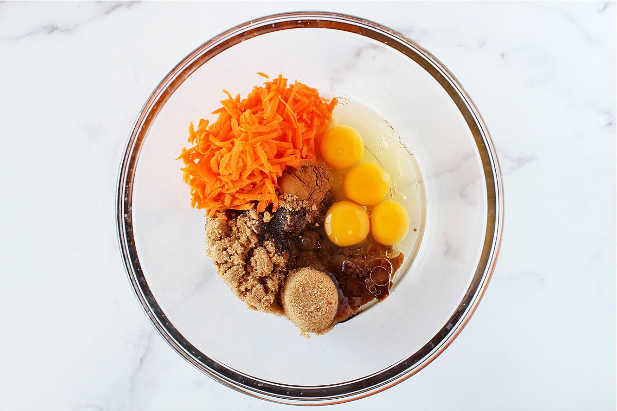 Shredded carrots, four eggs, golden monk fruit sweetener and oil in a glass mixing bowl.