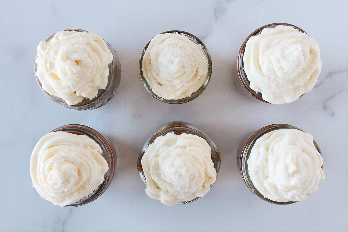 Six single serve carrot cake jars topped with cream cheese frosting and course sugar.