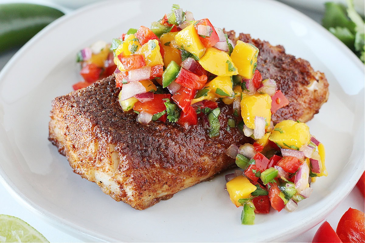 A blackened cod filet on a white plate topped with mango salsa.
