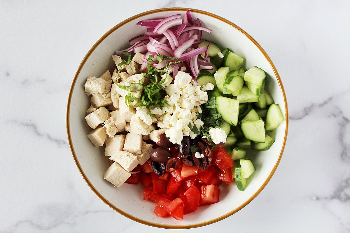 Feta cheese, basil, Kalamata olives, fresh veggies and diced chicken in a ceramic bowl.