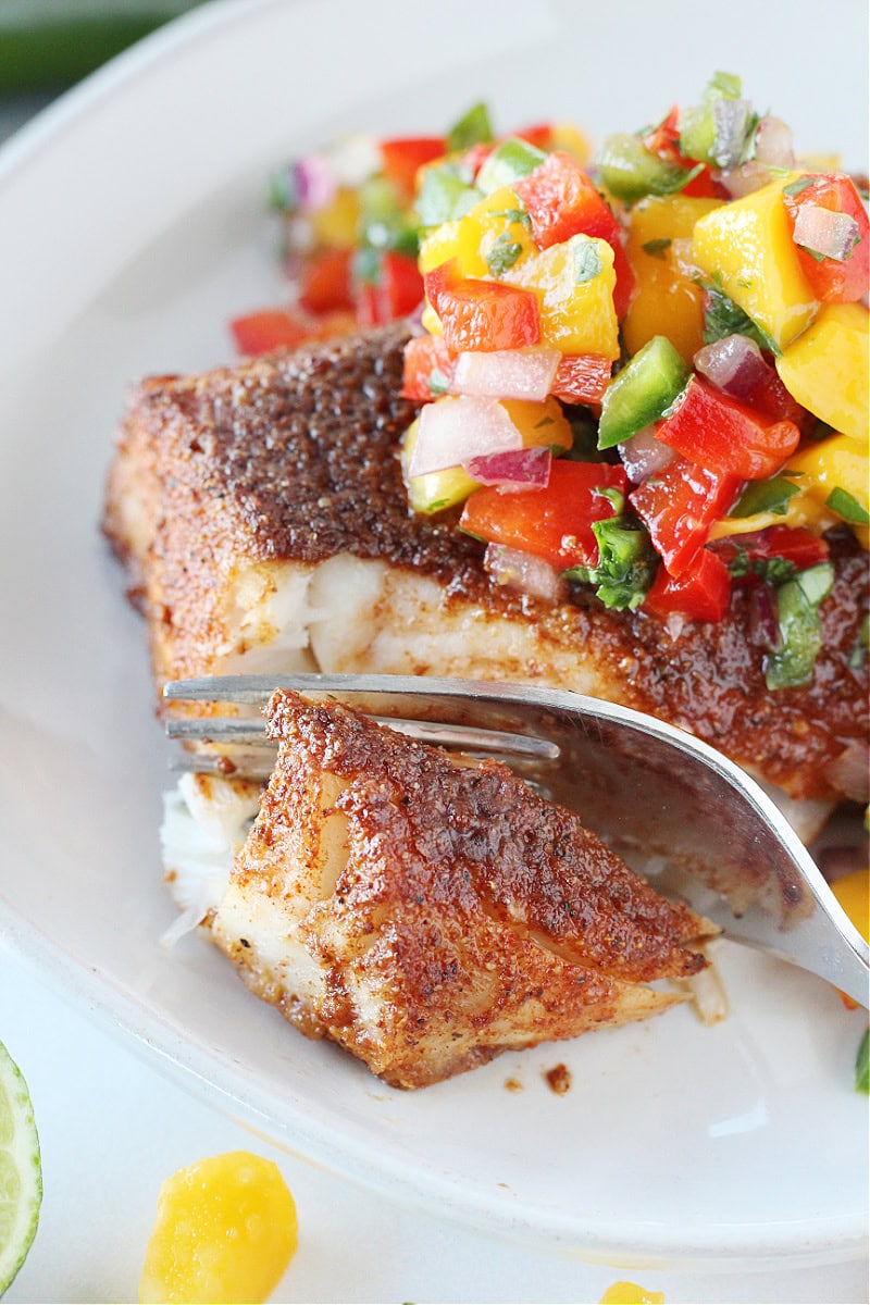 A fork slicing through a blackened cod filet topped with colorful mango salsa.