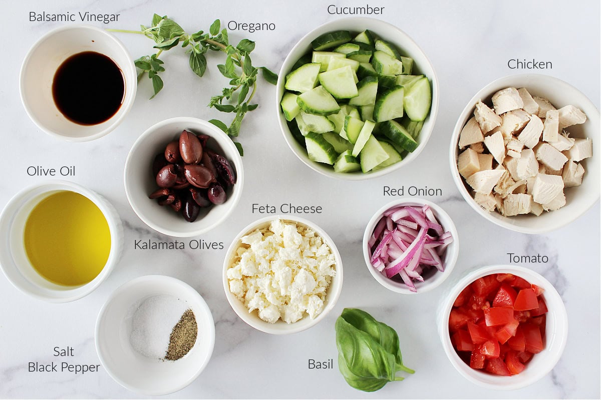 Labeled ingredients for grilled chicken Greek salad with homemade dressing on a white marble countertop.