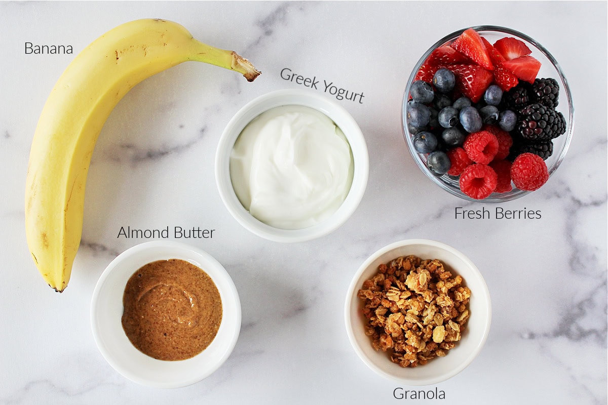 Labeled ingredients for a breakfast banana split on a white marble countertop.