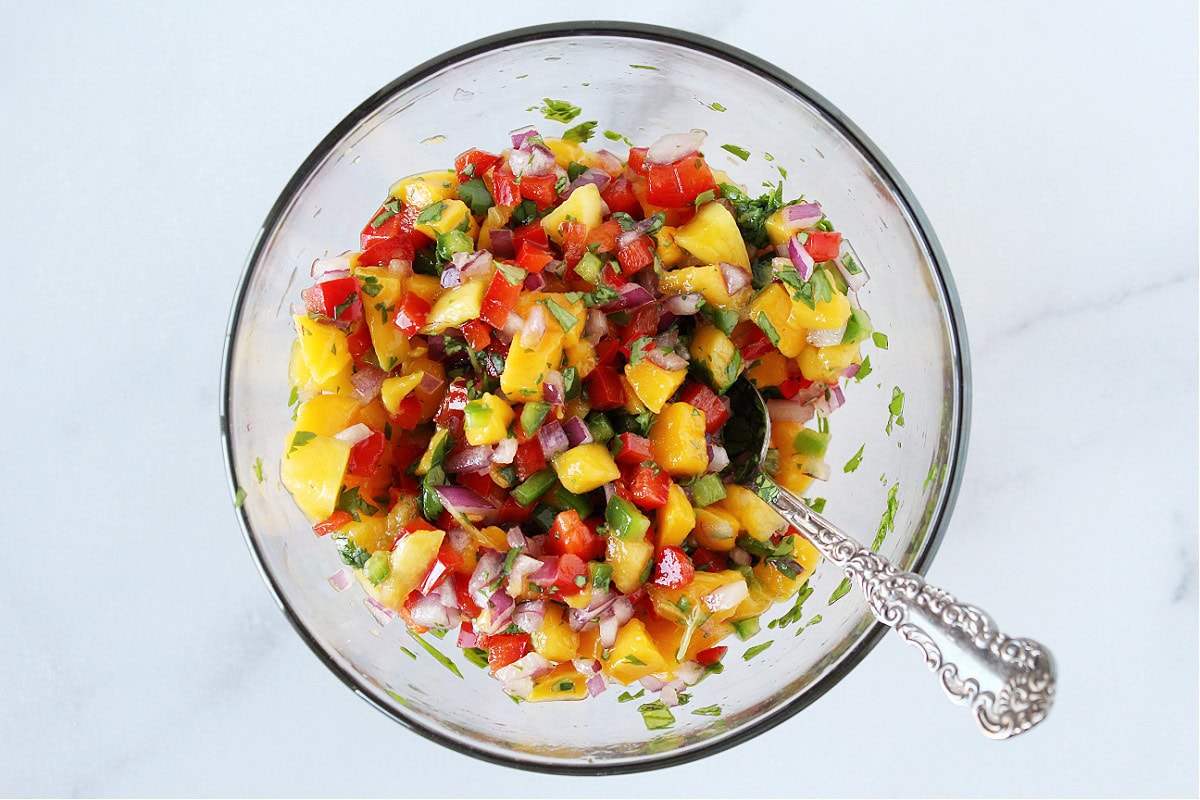 A spoon in a glass bowl of colorful mango salsa with red pepper and red onion.