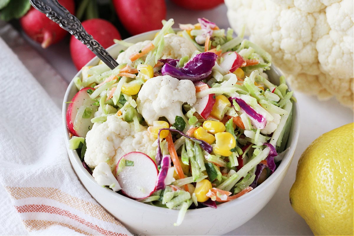Cauliflower florets, corn and sliced radish in a broccoli slaw salad in a bowl.