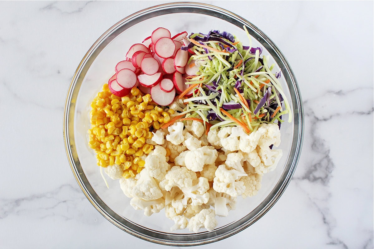 Sliced radish, corn, cauliflower florets and broccoli slaw in a glass bowl.