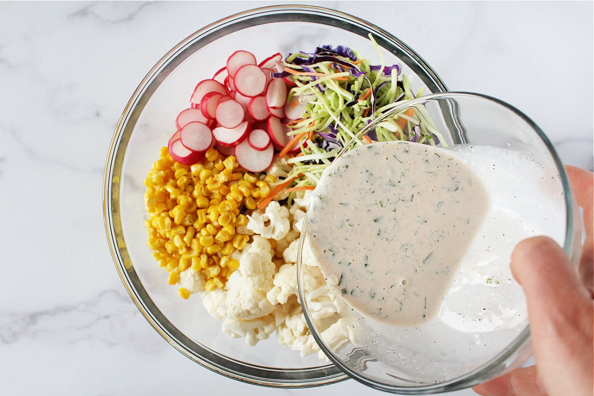 Pouring a mayo and basil dressing over fresh veggies in a glass bowl.