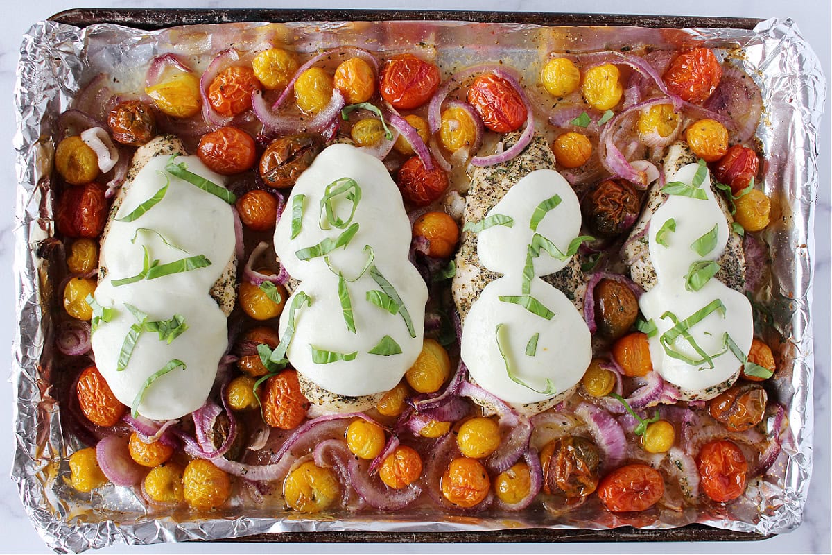 Four baked chicken breasts topped with mozzarella and basil with cherry tomatoes and red onion.