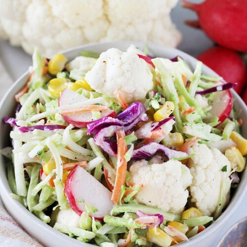 Cauliflower florets, radish, and corn in a broccoli slaw salad in a white bowl.