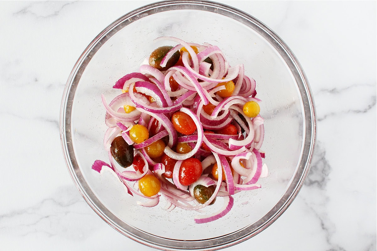 Sliced red onion and colorful cherry tomatoes coated in oil and seasonings in a bowl.