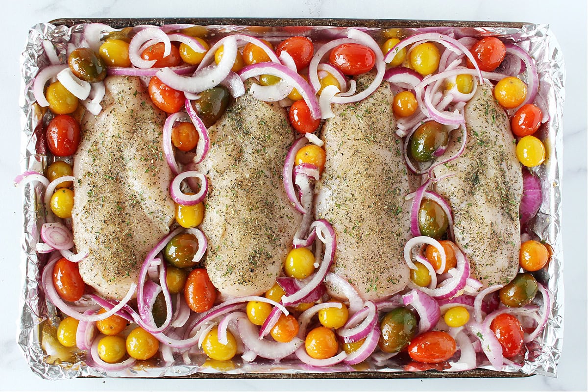 Four raw chicken breast topped with Italian seasoning surrounded by colorful tomatoes and red onion.
