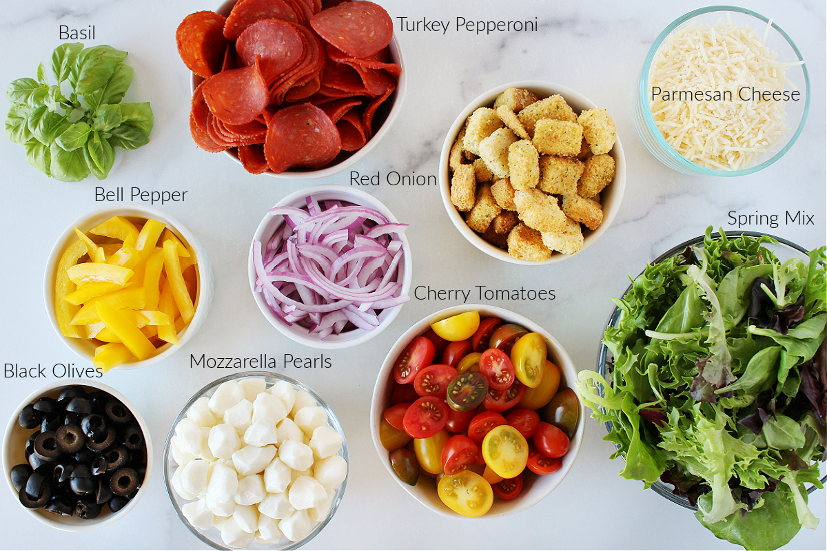 Labeled ingredients in bowls for pizza salad on a white marble countertop.