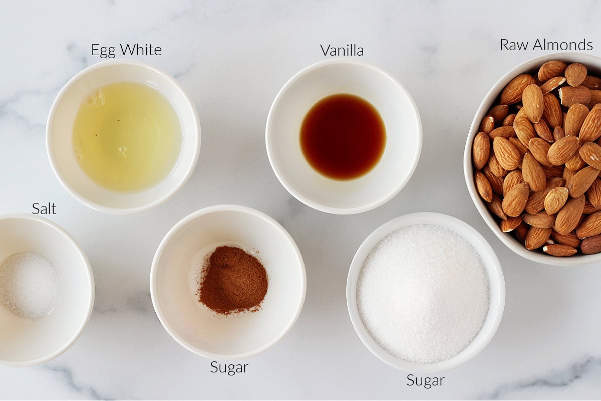 Labeled ingredients for candied almonds in bowls on a white marble countertop.