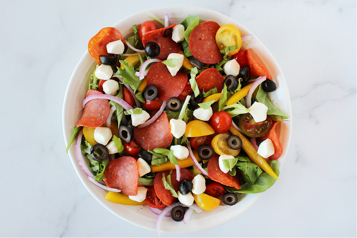 Mozzarella pearls, turkey pepperoni, black olives and veggies on mixed greens in a bowl.