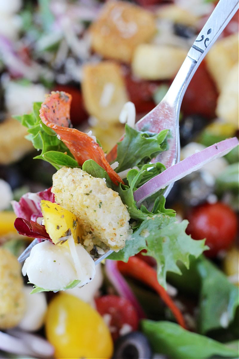Pizza salad ingredients like pepperoni, a crouton and veggies on a fork.