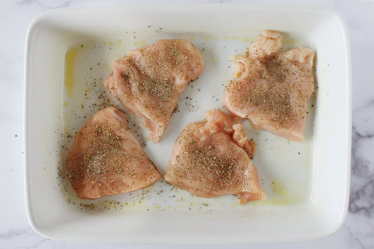 Four thin chicken breasts topped with Italian seasoning in a 9x13 ceramic dish.