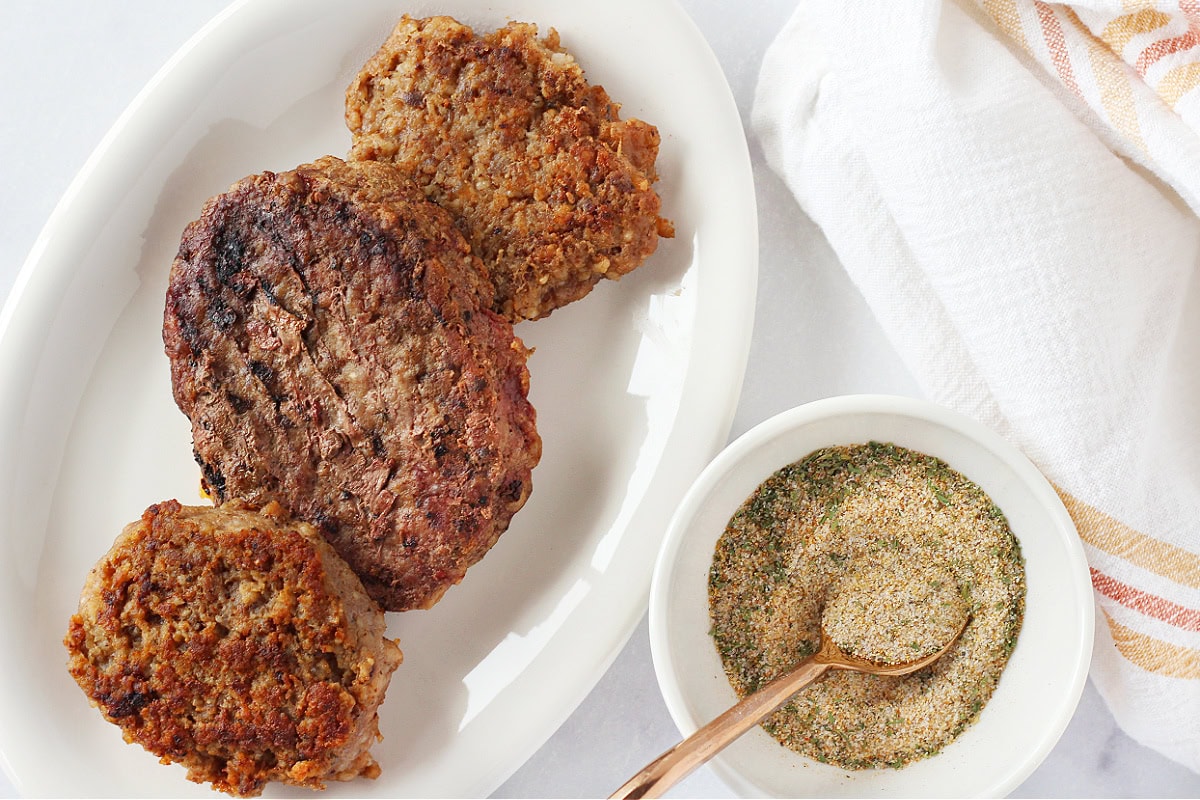 Three burgers on a white plate next to a bowl of homemade seasoning.