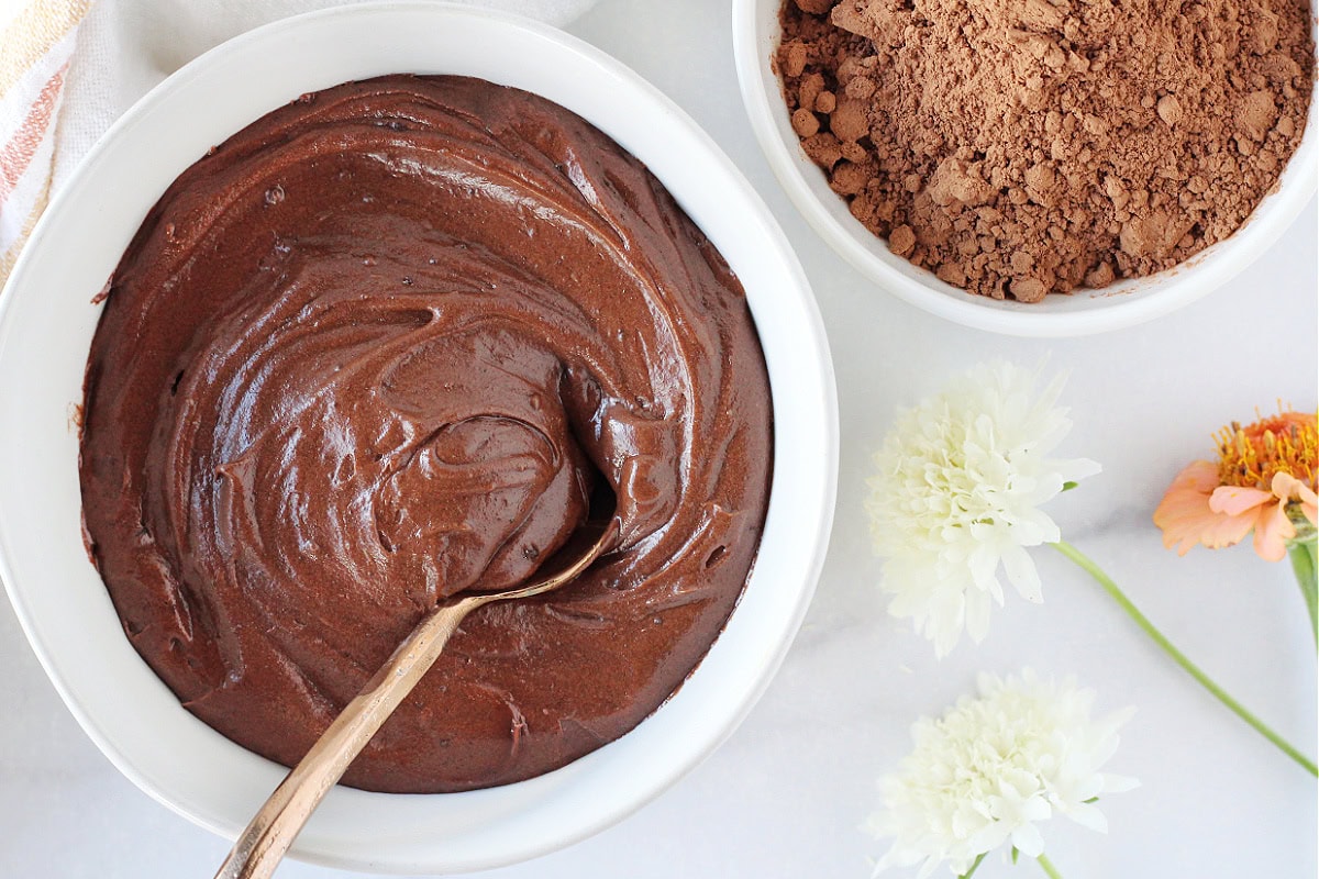 Chocolate frosting in a bowl with a spoon next to cocoa powder and flowers.