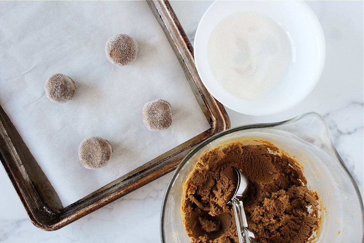 Cookie dough balls on a baking sheet, bowl of sugar, and dough in bowl.
