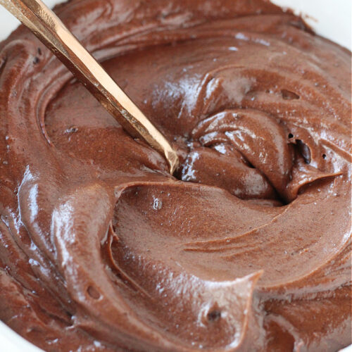 Chocolate frosting in a white bowl with a copper spoon.
